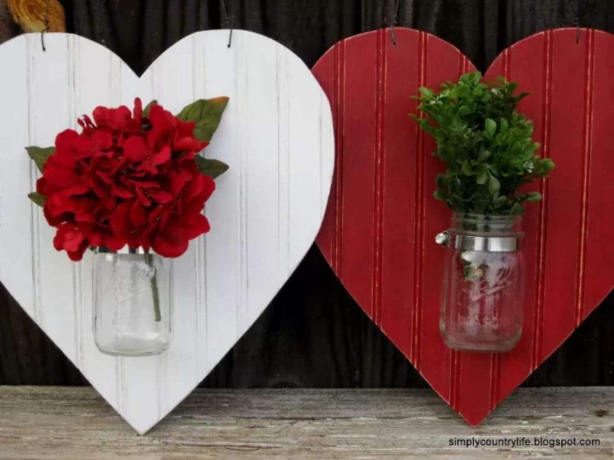 a white wooden heart is propped up next to a red wooden heart. Each heart has a glass mason jar stuck the front. inside the mason jar is a bunch of red flowers and green foliage